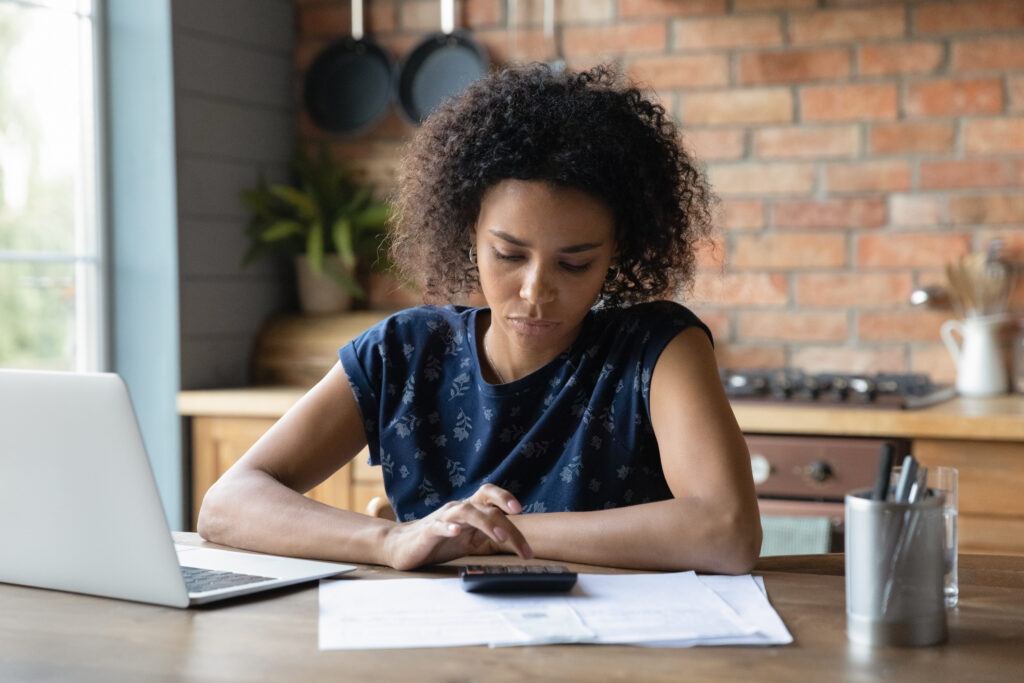 serious mixed race female using a calculator and a laptop in a kitchen with pencils and paper around her looking into how to improve her business credit