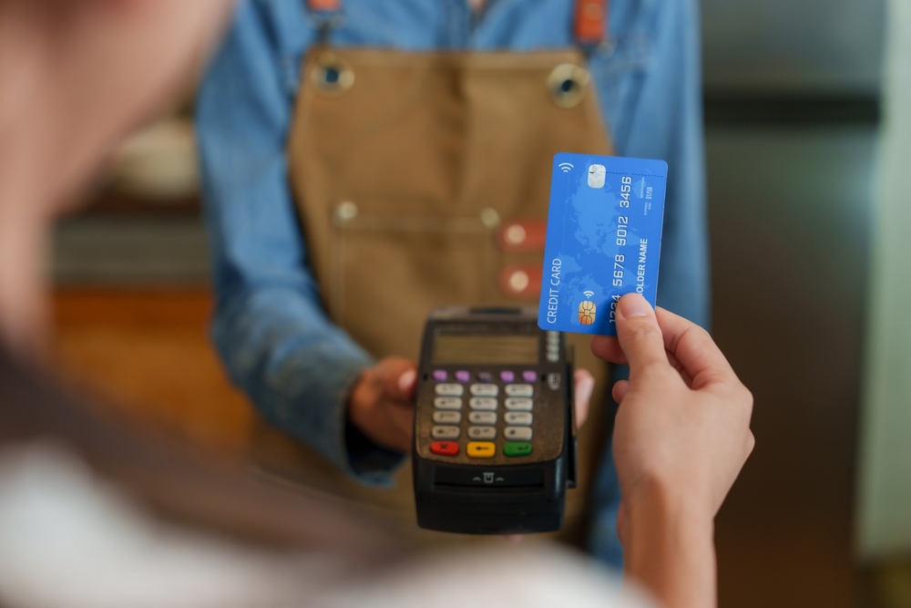 person with credit card in hand about to use on a  payment device held by a worker in a blue shirt and a brown overall cover