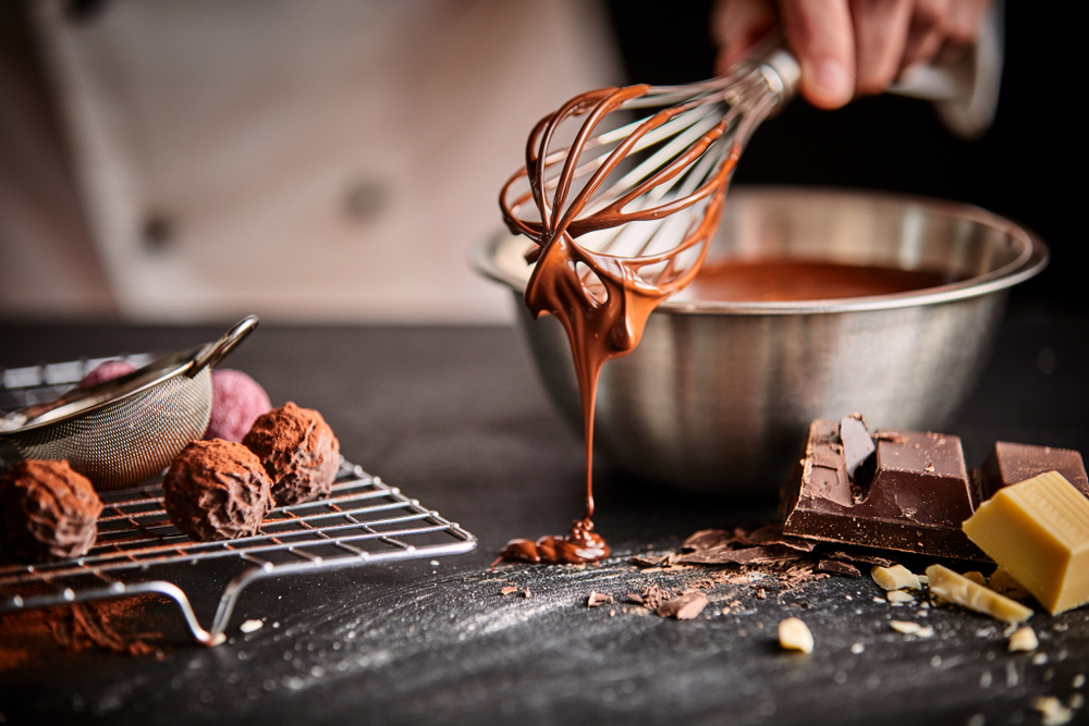 baker making cookies with a whisk dripping with mixture over a bowl with cookie balls on a tray