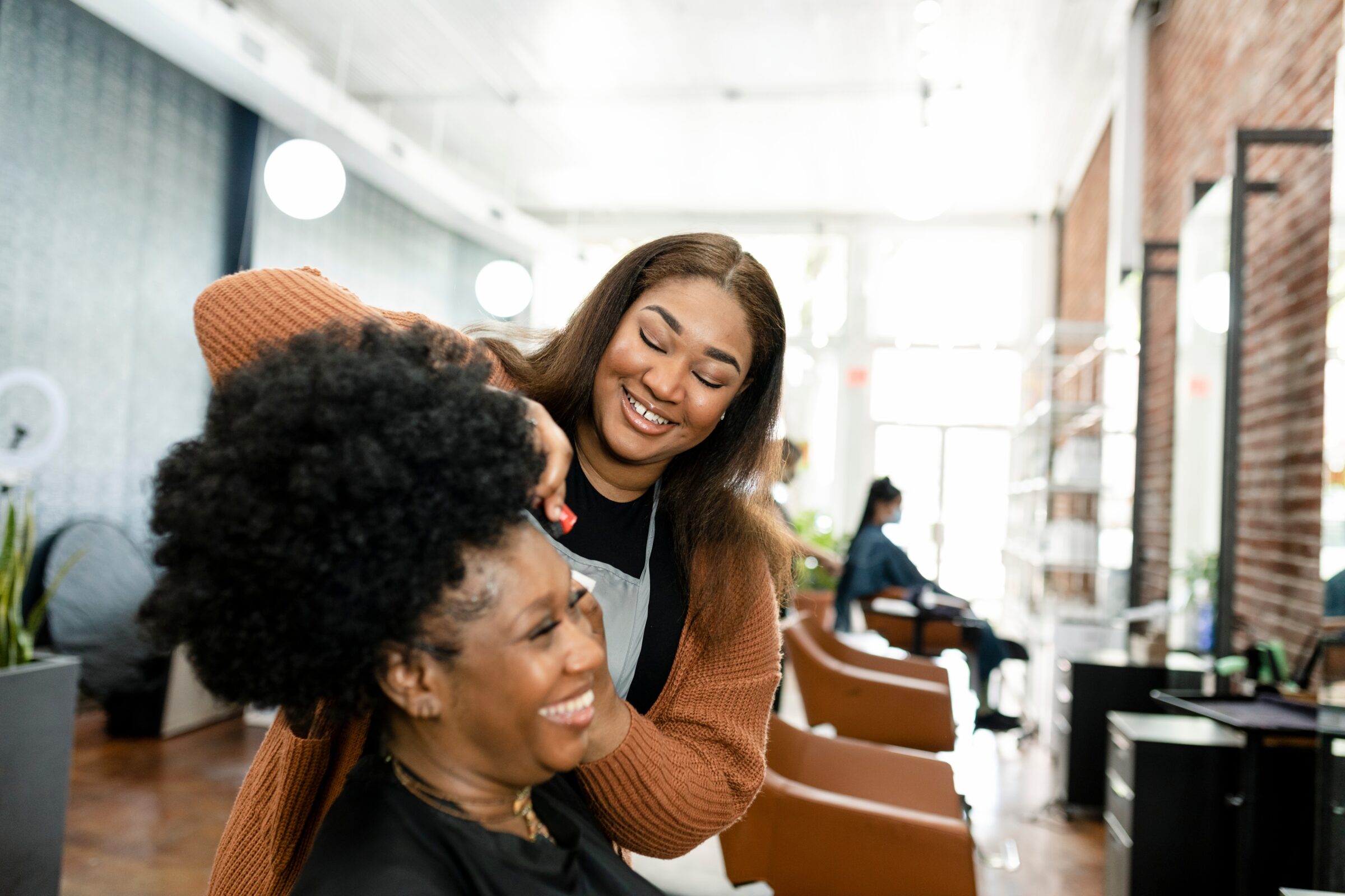 2 black women laughing while one women-owned business worker does the other's hair